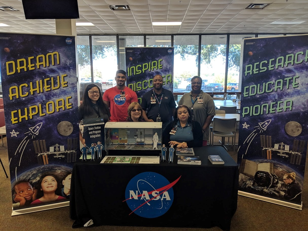 A group of 6 men and women stands behind an exhibit table about the International Space Station.